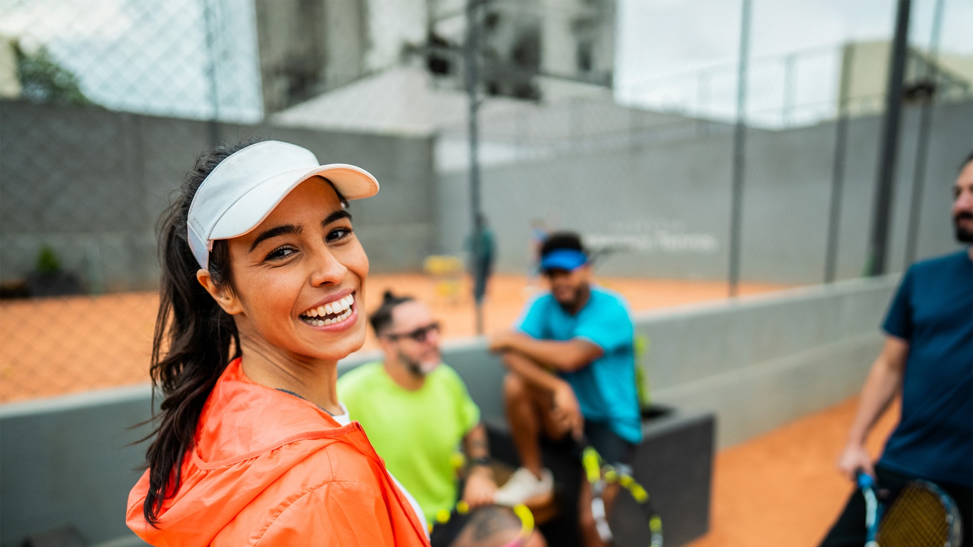 Smiling woman in a white visor and orange jacket at an outdoor tennis court, with players and a fenced court in the background