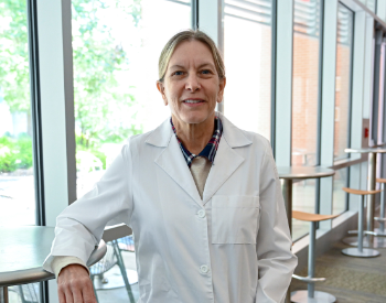 Standing women poses with one arm resting a table in her dental white coat.